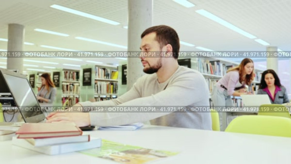 Focused guy student preparing for an exam on a laptop in the university library, looking for sources of information in textbooks. Стоковое видео, видеограф Яков Филимонов / Фотобанк Лори