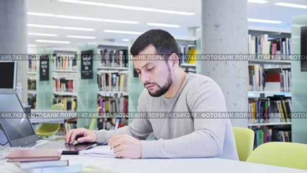 Focused guy student preparing for an exam on a laptop in the university library, looking for sources of information in textbooks. Стоковое видео, видеограф Яков Филимонов / Фотобанк Лори