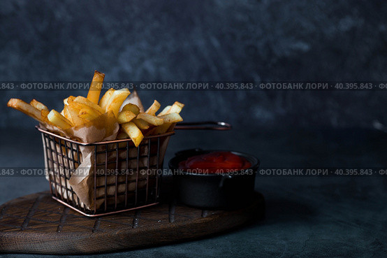 French fries in a metal basket with salt and ketchup on a stylish board on a dark background. Fried potatoes. The concept of fast food and junk food. Стоковое фото, фотограф Photojuli / Фотобанк Лори