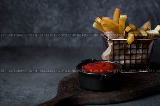 French fries in a metal basket with salt and ketchup on a stylish board on a dark background. Fried potatoes. The concept of fast food and junk food. Стоковое фото, фотограф Photojuli / Фотобанк Лори