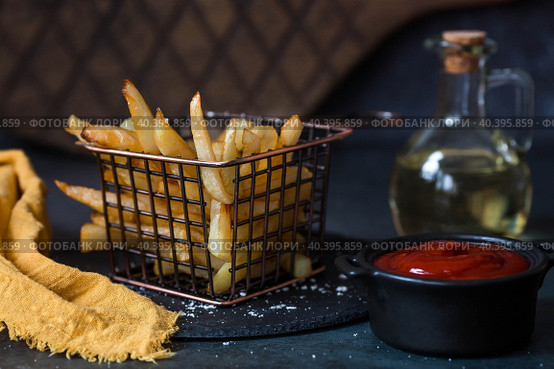 French fries in a metal basket with salt, ketchup and sunflower oil on a stylish board on a dark background. Fried potatoes. The concept of fast food and junk food. Стоковое фото, фотограф Photojuli / Фотобанк Лори