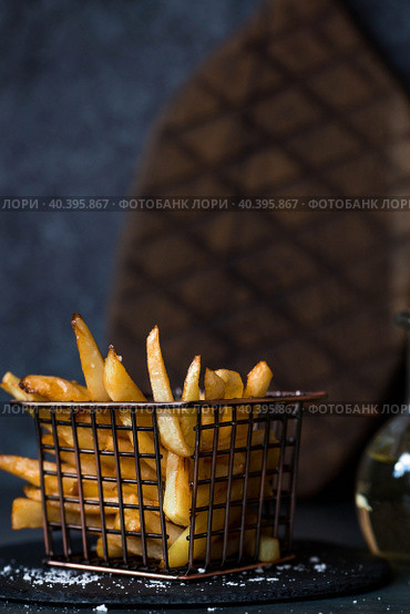 French fries in a metal basket with salt, ketchup and sunflower oil on a stylish board on a dark background. Fried potatoes. The concept of fast food and junk food. Стоковое фото, фотограф Photojuli / Фотобанк Лори