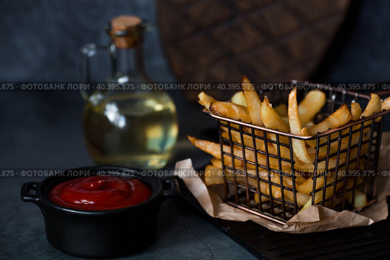 French fries in a metal basket with salt, ketchup and sunflower oil on a stylish board on a dark background. Fried potatoes. The concept of fast food and junk food. Стоковое фото, фотограф Photojuli / Фотобанк Лори
