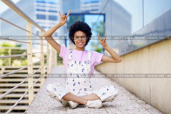 Full body of carefree African American female in casual outfit looking at camera with rock and roll gestures while sitting on street. Rebellious black woman showing horn signs. Стоковое фото, агентство Ingram Publishing / Фотобанк Лори