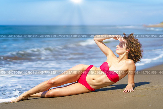 Full body of curly female in stylish bikini lying on sandy shore and covering eyes from bright sun. Gorgeous lady in bikini sunbathing on beach. Стоковое фото, агентство Ingram Publishing / Фотобанк Лори