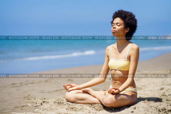 Full body of peaceful slim black female tourist in bikini sitting in Lotus pose on sandy seashore with closed eyes, while meditating during yoga session on sunny summer day. Fit young African American woman in Padmasana meditating on sandy beach. Стоковое фото, агентство Ingram Publishing / Фотобанк Лори