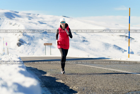 Full body of young female in red vest cap polarized sunglasses, looking at camera while jogging on asphalt road near snow against snowy mountain and blue sky in sunlight. Young woman enjoying running on road in winter clothes. Стоковое фото, агентство Ingram Publishing / Фотобанк Лори