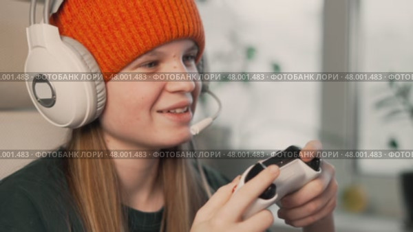 Girl enjoying video game in headphones with microphone, sitting comfortably at desk. Bright daylight, stylish tech gadgets and vibrant beanie create positive modern vibe. Стоковое видео, видеограф Енваев Андрей / Фотобанк Лори