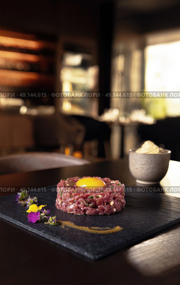 Gourmet beef tartare with raw egg yolk, beautifully plated with edible flowers and sauce, served on black slate in upscale restaurant setting. Стоковое AI-изображение, AI-художник Катерина Бондарец / Фотобанк Лори
