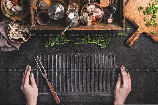 Grill or BBQ food background. Woman hold empty cast iron grill griddle and meat fork on roast rustic kitchen table. Wooden box with seasonings, oils and other kitchen utensils.Top view. Place for text. Стоковое фото, агентство Ingram Publishing / Фотобанк Лори