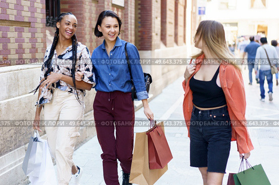 Group of cheerful multiracial female friends with shopping bags of purchases looking at each other while strolling on sidewalk near building in city. Smiling multiethnic women walking on street after shopping. Стоковое фото, агентство Ingram Publishing / Фотобанк Лори