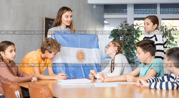 Group of curious preteen learners and young female teacher with flag of Argentina in classroom of academy. Стоковое фото, фотограф Яков Филимонов / Фотобанк Лори