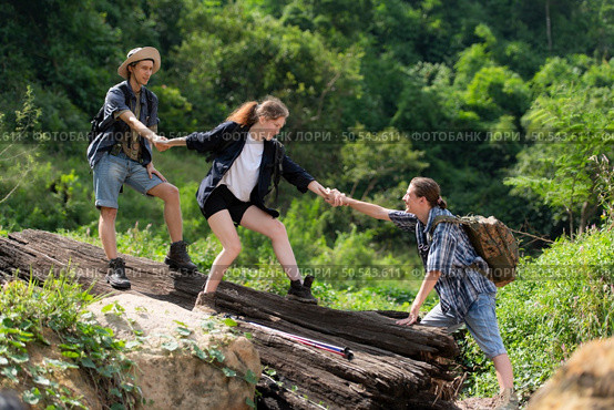 Group of friend on a sunny day in the forest Young group hiker with backpack and trekking poles helping to pull each other up to stand on the log. Стоковое фото, агентство Ingram Publishing / Фотобанк Лори