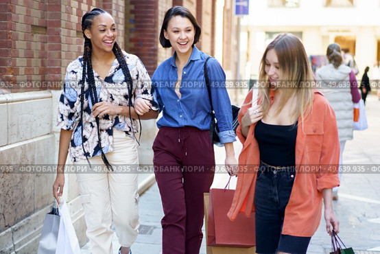 Group of optimistic multiethnic female friends with shopping bags strolling together on paved street near building after shopping in city. Content multiracial women with shopping bags. Стоковое фото, агентство Ingram Publishing / Фотобанк Лори