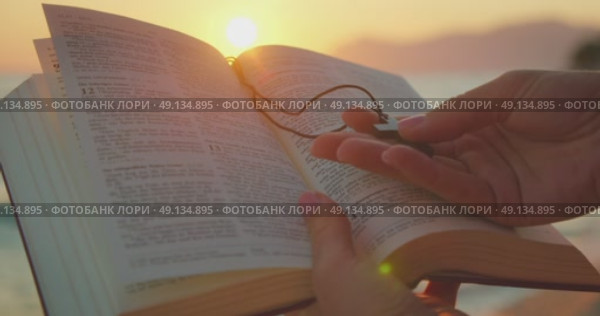Hands hold an open Bible and a cross pendant at the beach during a sunset as the light shines over the pages. Стоковое видео, видеограф Igor Tichonow / Фотобанк Лори