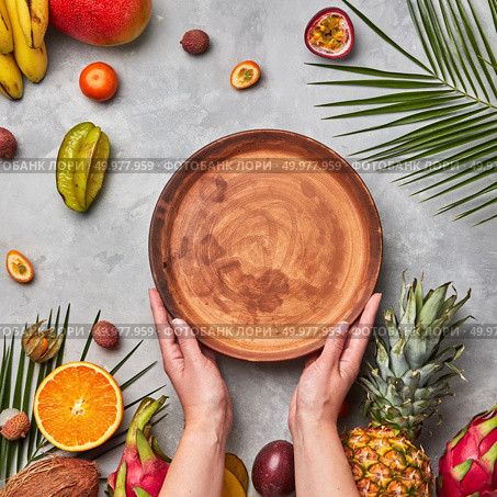 Hands of a woman holding a brown empty plate around a different tropical ripe fruit, coconut, pineapple, physalis, banana on a gray concrete background with copy space. Flat lay. Ripe tropical fruits, coconut, mango, carambola, lychee and palm leaves on a gray concrete background. The woman’s hands hold an empty brown round plate with a copy of the space. Flat lay. Стоковое фото, агентство Ingram Publishing / Фотобанк Лори