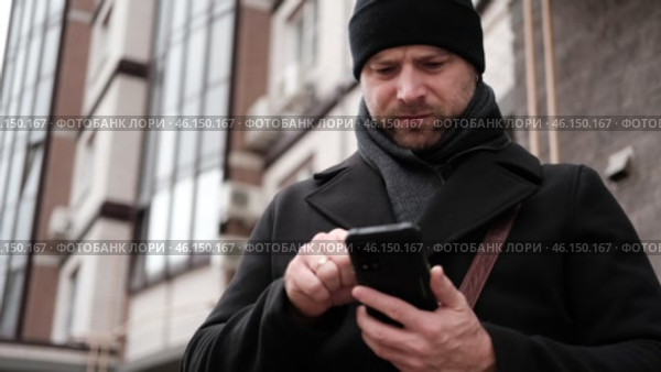 Handsome man wearing black coat and brown leather bag walking down on the city with mobile phone in hand. Stylish bearded middle-aged male using smartphone at urban street. Businessman going on job. Стоковое видео, видеограф Марина Демидюк / Фотобанк Лори