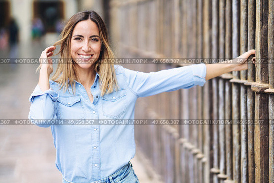 Happy adult female in stylish blue shirt looking at camera with smile and touching hair while grasping metal fence on blurred background of city street. Cheerful woman touching hair near fence. Стоковое фото, агентство Ingram Publishing / Фотобанк Лори