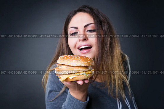 Happy beautiful young plus size model posing with hamburger on a gray studio background, xxl woman eating burger. Стоковое фото, фотограф Станислав Тарко / Фотобанк Лори