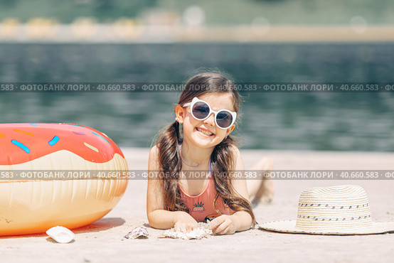 Happy cute smiling little girl in sunglasses lying on the sand on a beach in a swimsuit near a straw hat. Стоковое фото, фотограф Ирина Михайличенко / Фотобанк Лори