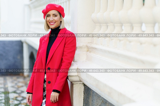 Happy female in fashionable red clothes with beret looking at camera with smile while standing near white fencing in city. Cheerful woman in stylish outfit near fence. Стоковое фото, агентство Ingram Publishing / Фотобанк Лори