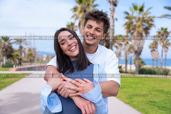 Happy young couple hugging and smiling on palm-lined seaside promenade. Стоковое фото, фотограф Raquel Corcoles Mendez / Фотобанк Лори