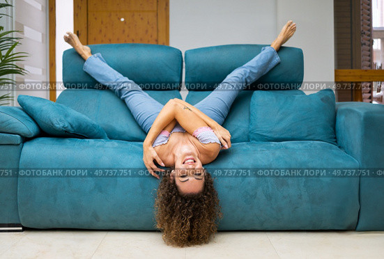Happy young female in jeans and top with curly hair lying upside down on sofa in living room and having fun at home while enjoying. Cheerful woman lying upside down on sofa at home. Стоковое фото, агентство Ingram Publishing / Фотобанк Лори