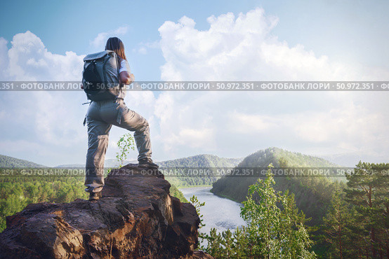 Hiker standing on rocky ledge overlooking river and forest landscape during daytime under blue sky . A female traveler with a backpack on the Mana River and the Manskaya Loop in Siberia, Russia Krasnoyarsk. Стоковое фото, фотограф Владимир Разгуляев / Фотобанк Лори