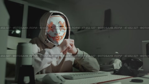 Hooded individual in light jacket types on keyboard wearing symbolic glowing mask. Captures energy of digital movement and hacker as modern visual activist. Ethical hacker cybersecurity concept. Стоковое видео, видеограф Енваев Андрей / Фотобанк Лори