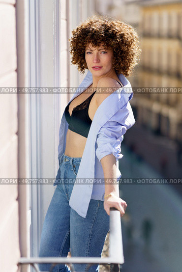Hot female with brown curly hair in unbuttoned shirt and black bra leaning on railing of balcony looking at camera. Attractive woman with unbuttoned shirt standing on balcony. Стоковое фото, агентство Ingram Publishing / Фотобанк Лори