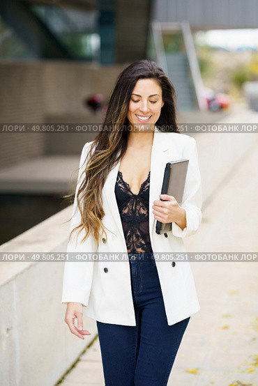 In a busy urban area, a stylish businesswoman in a chic outfit strides confidently down the crowded sidewalk, holding her sleek tablet, embodying modern elegance and selfassuredness. A Stylish and Modern Businesswoman Walking Confidently Outdoors while Holding a Tablet in Her Hands. Стоковое фото, агентство Ingram Publishing / Фотобанк Лори