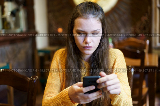 In a cozy and warm cafe setting, a young woman with long flowing hair is sitting comfortably, wearing a bright yellow sweater, completely focused on her smartphone, absorbed in her digital world. A young woman is deeply engrossed in her smartphone while sitting inside a cozy cafe. Стоковое фото, агентство Ingram Publishing / Фотобанк Лори
