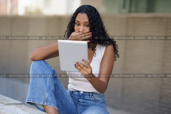 In a lively outdoor scene, a young woman shows surprise, hands covering her mouth in disbelief as she gazes at her tablet, overwhelmed by emotions that capture her attention and curiosity. A Surprised Young Woman Engaging with a Tablet While Sitting in a Beautiful Outdoor Setting. Стоковое фото, агентство Ingram Publishing / Фотобанк Лори