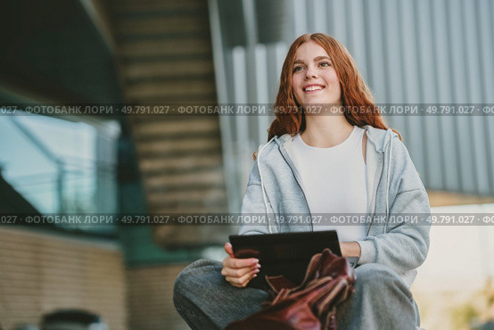 In a lively urban environment, a stylish young woman is seated outside, joyfully engaged with her tablet on this stunning sunny day, fully relishing every moment of her vibrant life experience. A Young Woman Delightfully Engaging with Her Tablet in a Vibrant Urban Outdoor Environment. Стоковое фото, агентство Ingram Publishing / Фотобанк Лори