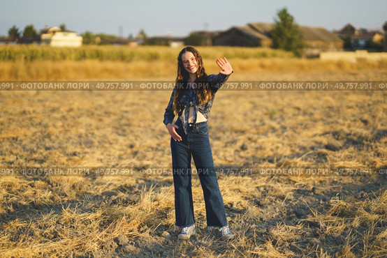 In a sunlit golden field, a young woman beams with joy as she waves cheerfully, dressed in a trendy denim outfit that perfectly embodies a warm and relaxed vibe, radiating happiness and freedom. A Young Woman Dressed in a Stylish Denim Outfit Waves Cheerfully in a Beautiful Golden Field. Стоковое фото, агентство Ingram Publishing / Фотобанк Лори