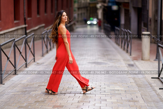 In a vibrant city, a fashionable woman confidently walks down a tranquil street in a striking red jumpsuit, showcasing her unique style and elegance, captivating everyone with her grace and poise. A Stylish Woman Dressed in a Bold Red Jumpsuit is Walking Alone in an Urban Setting. Стоковое фото, агентство Ingram Publishing / Фотобанк Лори