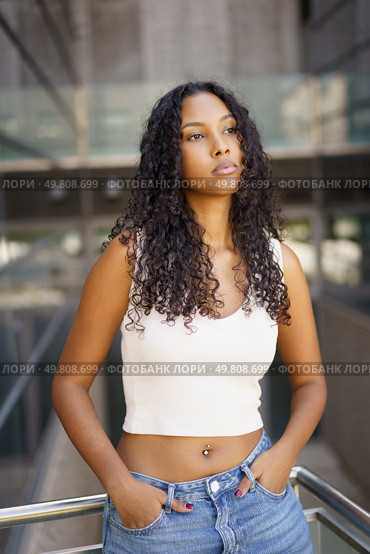 In a vibrant urban setting, a confident young woman with curly hair embodies modern youth culture through her distinct style and charm, reflecting a contemporary vibe that is engaging and captivating. A Confident Young Woman Posing Gracefully in a Modern Urban Setting Under Bright Daylight. Стоковое фото, агентство Ingram Publishing / Фотобанк Лори