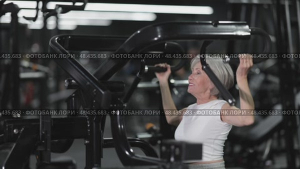 In gym, senior woman uses lat pulldowm machine to strengthen arm muscles. Стоковое видео, видеограф Яков Филимонов / Фотобанк Лори