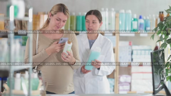 Interested pregnant woman using smartphone to check ingredients of organic shampoo at drugstore, while smiling professional female pharmacist in background suggesting different pregnancy-safe option. Стоковое видео, видеограф Яков Филимонов / Фотобанк Лори