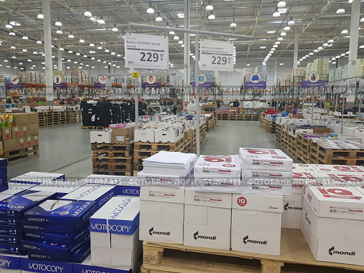 Interior of a large wholesale hypermarket. A spacious sales area with a high ceiling and powerful lighting is filled with rows of pallets with goods. In the center of the frame are packs of A4 office paper of different brands, in particular VOTOCOPY and Mondi IQ Economy. The paper is packed in boxes and stacks on wooden pallets. The price tags indicate the cost - 229.99 rubles for a pack of 500 sheets. In the background, you can see racks with other categories of goods, such as food, household chemicals, hygiene products and office supplies. The atmosphere of the photo reflects a large warehouse-shop with self-service. (2019 год). Редакционное фото, фотограф Анна Кривицкая / Фотобанк Лори