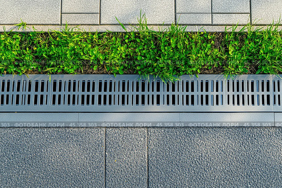 Iron grate of a drainage system for storm water drainage from a pedestrian sidewalk near a green lawn. Drain system installation. City metal grate and concrete street drain on the sidewalk of a road. Стоковое AI-изображение, AI-художник Марина Демидюк / Фотобанк Лори