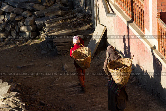 Jharkot, Nepal - November 18, 2015: Nepalese women carry on their back baskets for cargo along the street in the village in Nepal. Редакционное фото, фотограф VittoriaChe / Фотобанк Лори