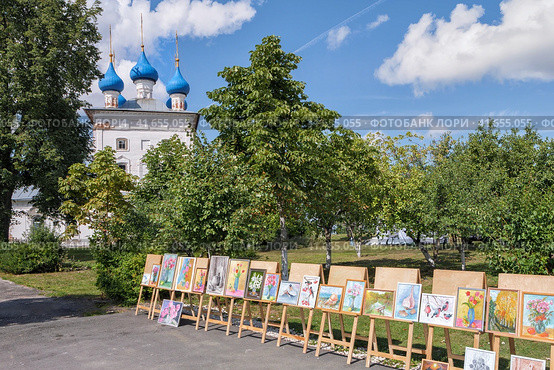 Клязьминский городок (Стародуб), Россия. Выставка рисунков деревенских детей-художников на фоне зеленого сада, старинного храма с голубыми куполами и синего неба с белыми облаками. (2023 год). Стоковое фото, фотограф Сергей Никифоров-Гордеев / Фотобанк Лори