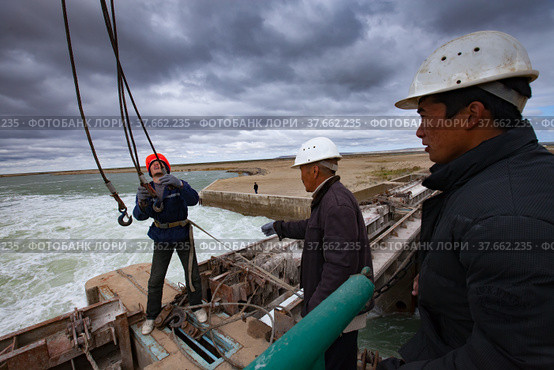 Kok-aral, Kazakhstan - May 03, 2012: Small Aral Sea Kok-aral dam. Hydraulic engineers prepair to lift water shutter weights. Crane's steel rope with hooks. Stormy sky and turbulent water. Редакционное фото, фотограф Alexey Rezvykh / Фотобанк Лори
