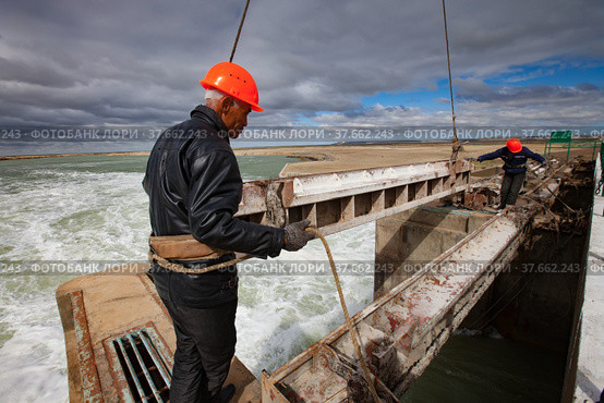 Kok-aral, Kazakhstan - May 03, 2012: Small Aral Sea Kok-aral dam. Two workers in orange helmets. Lifts water shutter weighting agent (steel girder) by crane. Редакционное фото, фотограф Alexey Rezvykh / Фотобанк Лори