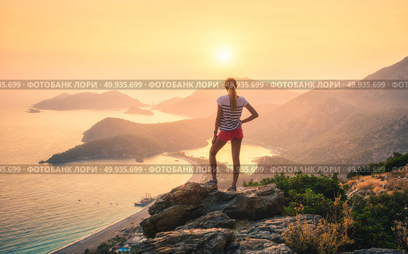 Landscape with woman, sea, mountain ridges and orange sky. Young woman standing on the top of rock and looking at the seashore and mountains at colorful sunset in summer. Landscape with girl, sea, mountain ridges and orange sunlight. Travel. Oludeniz, Turkey. Стоковое фото, агентство Ingram Publishing / Фотобанк Лори