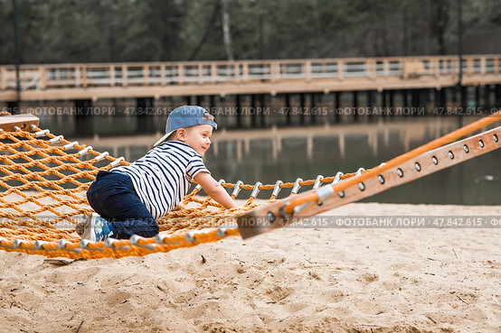 Lifestyle. A cute little boy is sitting in a hammock on a modern outdoor playground. The concept of family entertainment and leisure. Стоковое фото, фотограф Photojuli / Фотобанк Лори
