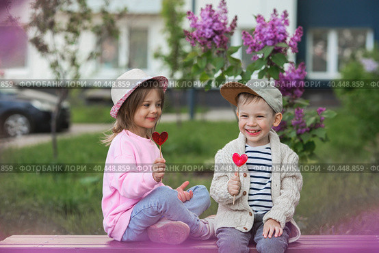 Lifestyle. Little beautiful children are playing on a bench with candy lollipops on a stick in the shape of a heart. The concept of childhood friendship and first love. Стоковое фото, фотограф Photojuli / Фотобанк Лори