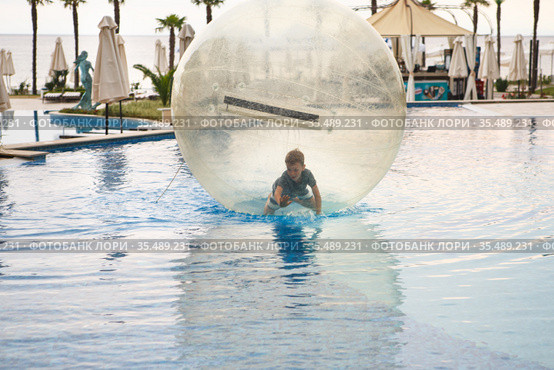 Little boy have fun inside big plastic balloon on the water of swimming pool on the summer resort. Kid inside big inflatable transparent ball running and having fun. Стоковое фото, фотограф Nataliia Zhekova / Фотобанк Лори