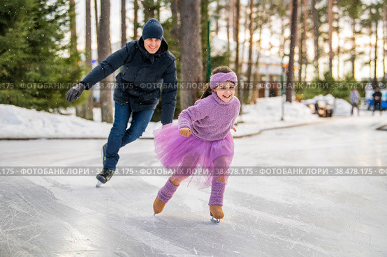 Little girl in pink sweater and full skirt rides on sunny winter day on an outdoor ice rink in park Девочка в красивом сиреневом свитере и юбке из тафты катается с папой на коньках. Стоковое фото, фотограф Мария Мороз / Фотобанк Лори